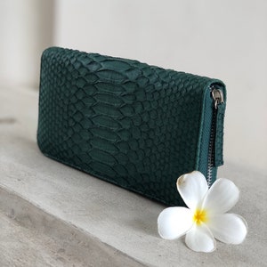 以下が含まれることがあります： ジッパー付きの濃い緑色のヘビ革財布。財布は薄い灰色の表面に白い花と一緒に置かれています。