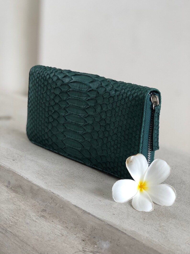 以下が含まれることがあります： ジッパー付きの濃い緑色のヘビ革財布。財布は薄い灰色の表面に白い花と一緒に置かれています。