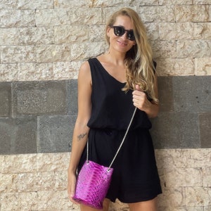 Op de afbeelding: Een vrouw in een zwarte, mouwloze jurk en zonnebril houdt een klein roze slangleren handtasje vast met een zilveren kettingband.