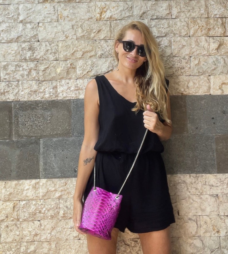 Op de afbeelding: Een vrouw in een zwarte, mouwloze jurk en zonnebril houdt een klein roze slangleren handtasje vast met een zilveren kettingband.