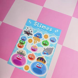 May include: A sheet of colourful stickers featuring various cartoon slimes with crowns, wings, and coins. The stickers are on a blue background with the word "Slimes" and star accents. The background is a pink and white chequered pattern.
