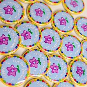 Pode incluir: Uma coleção de botões redondos e coloridos. Cada botão apresenta uma flor roxa em forma de estrela sobre um fundo azul claro, com as palavras "Certified Stardew Addict" em um padrão circular. Os botões têm uma borda amarela.