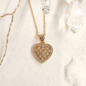 May include: A gold necklace featuring a heart-shaped pendant. The pendant is crafted from a delicate, openwork design and is embellished with small, clear stones.
