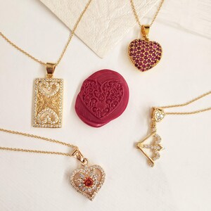 Puede incluir: Cuatro collares de oro con diferentes colgantes en forma de corazón. Un colgante es un corazón rojo con pequeñas piedras, otro es un corazón con un diseño floral, otro es un corazón con una sola piedra roja y el último es una flecha con un corazón en el extremo.