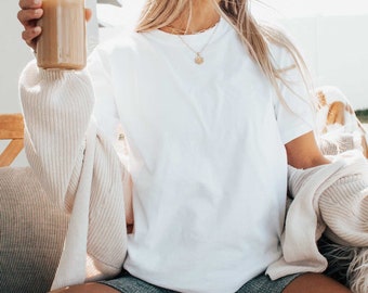 May include: A woman wearing a white t-shirt, gray shorts, and a cream cardigan sits on a fluffy white chair. She is holding a glass of iced coffee and wearing sunglasses.
