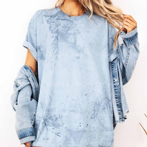 May include: Light blue t-shirt with a textured design and rolled-up sleeves. A light blue denim jacket is draped over the shoulders. The shirt has a faded, tie-dye appearance, creating a casual, relaxed look.