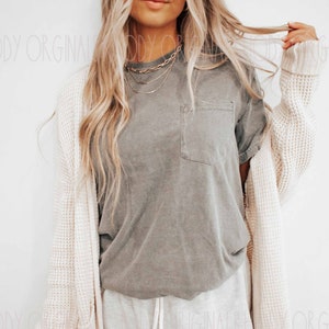 May include: A woman wearing a gray t-shirt with a pocket, a white cardigan, and white sweatpants. She is wearing a layered gold necklace.