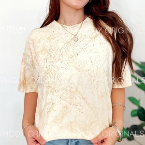 Pode incluir: Uma mulher est&aacute; vestindo uma camiseta tie-dye cor creme com um padr&atilde;o marrom e branco. Ela est&aacute; vestindo jeans azul e um colar de ouro com um pequeno pingente.