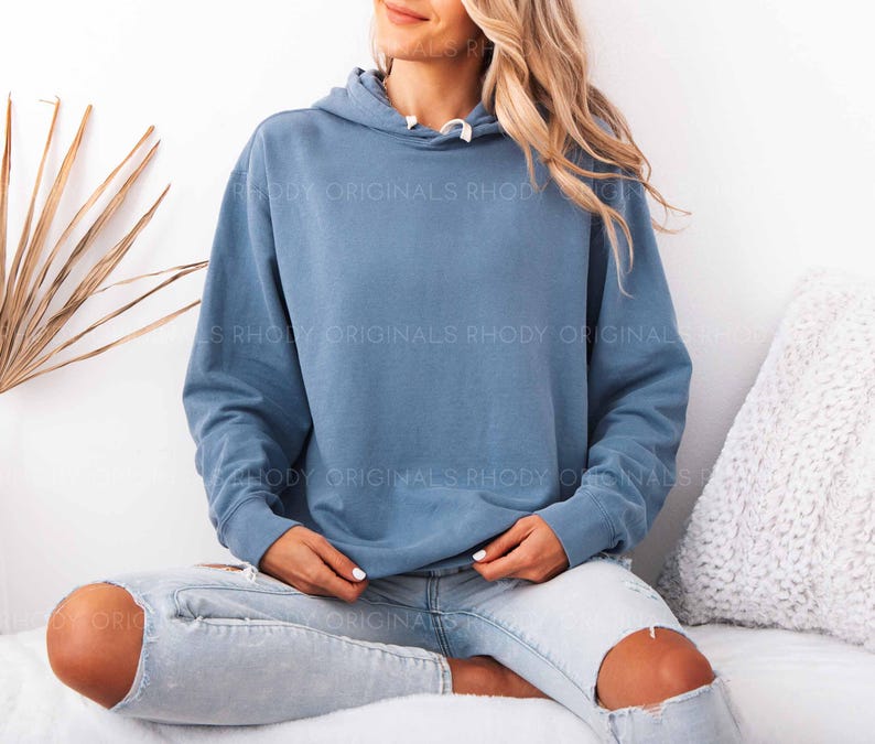 May include: A blue hoodie with a drawstring hood. The hoodie is worn with light blue jeans with ripped knees. The text "RHODY ORIGINALS" is printed on the hoodie. The person is sitting on a white surface.