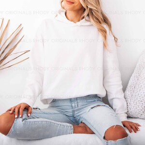 May include: A white hooded sweatshirt with long sleeves. The model is wearing light blue ripped jeans. The image is set against a white background with a dried palm leaf and a decorative pillow.