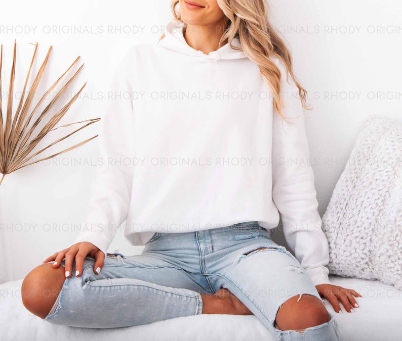 May include: A white hooded sweatshirt with long sleeves. The model is wearing light blue ripped jeans. The image is set against a white background with a dried palm leaf and a decorative pillow.