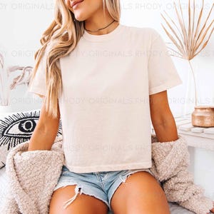 May include: A woman wearing a white cropped t-shirt with short sleeves and denim shorts. She is sitting on a white bed with a white throw blanket and a black and white eye pillow.