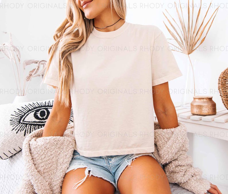 May include: A woman wearing a white cropped t-shirt with short sleeves and denim shorts. She is sitting on a white bed with a white throw blanket and a black and white eye pillow.