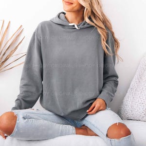 May include: A gray hoodie with a drawstring hood and a white drawstring. The hoodie is worn with light blue jeans with ripped knees. The text "RHODY ORIGINALS" is printed on the hoodie.