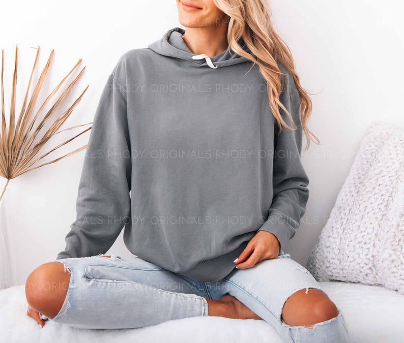May include: A gray hoodie with a drawstring hood and a white drawstring. The hoodie is worn with light blue jeans with ripped knees. The text "RHODY ORIGINALS" is printed on the hoodie.