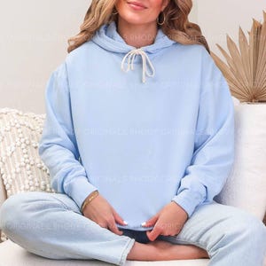 May include: A light blue hoodie with a drawstring, paired with light blue jeans. The hoodie has a kangaroo pocket and a relaxed fit. The model is wearing a gold bracelet and hoop earrings. The background includes a white couch and a dried palm leaf.