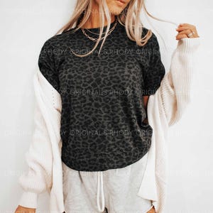 K&ouml;nnte beinhalten: Ein dunkelgraues T-Shirt mit Leopardenmuster und kurzen &Auml;rmeln. Das Shirt wird mit hellgrauen Jogginghosen und einer cremefarbenen Strickjacke kombiniert. Goldene Halsketten sind sichtbar.