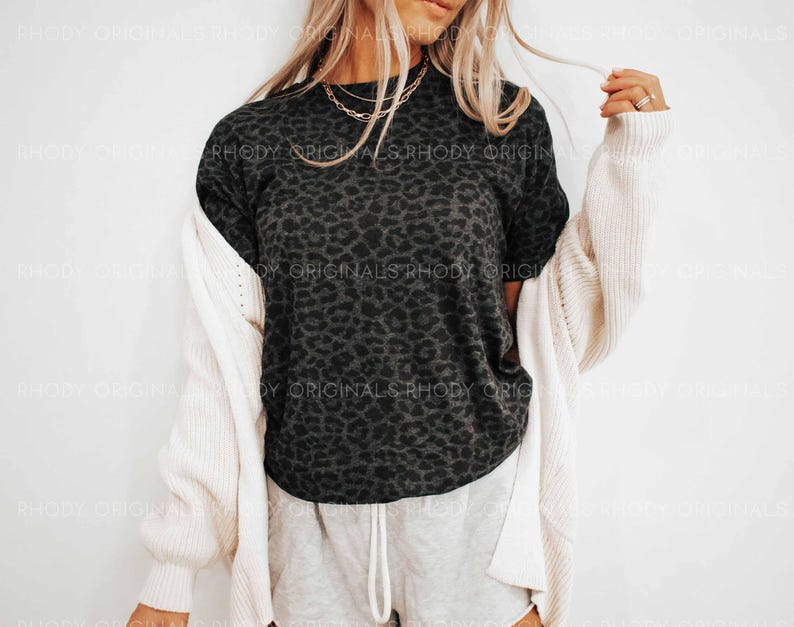 K&ouml;nnte beinhalten: Ein dunkelgraues T-Shirt mit Leopardenmuster und kurzen &Auml;rmeln. Das Shirt wird mit hellgrauen Jogginghosen und einer cremefarbenen Strickjacke kombiniert. Goldene Halsketten sind sichtbar.