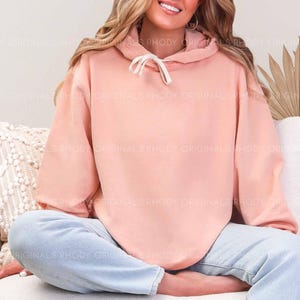 May include: A light peach-colored hoodie with a drawstring, paired with light blue jeans. The hoodie has a relaxed fit and is made of a soft material. The person is sitting on a white couch.