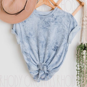 May include: A light blue tie-dye t-shirt with a knotted front. The shirt is hanging on a wooden hanger against a white wall.