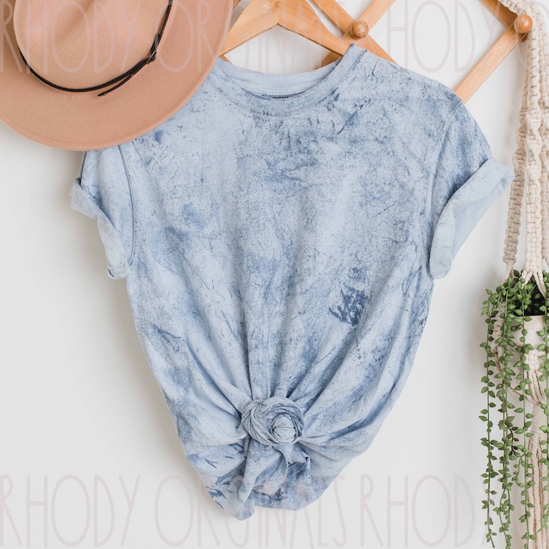 May include: A light blue tie-dye t-shirt with a knotted front. The shirt is hanging on a wooden hanger against a white wall.