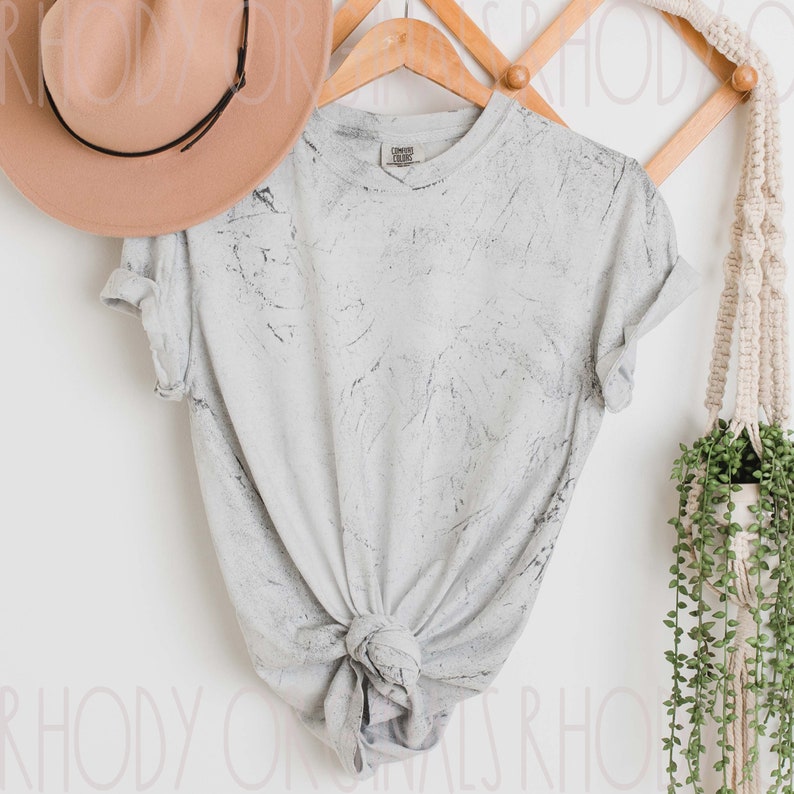 May include: A white t-shirt with a gray marble pattern. The t-shirt is hanging on a wooden hanger with a brown hat on top of it.