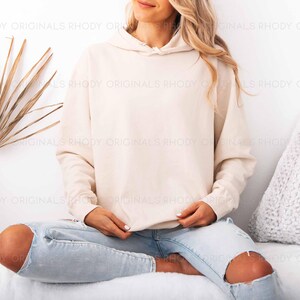 May include: A cream-colored hoodie with a drawstring hood. The model is wearing light blue ripped jeans. The background includes a white wall, a dried palm leaf, and a white pillow. The hoodie is a neutral color.