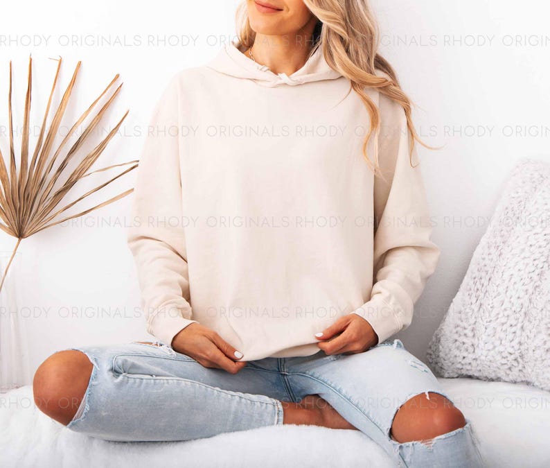 May include: A cream-colored hoodie with a drawstring hood. The model is wearing light blue ripped jeans. The background includes a white wall, a dried palm leaf, and a white pillow. The hoodie is a neutral color.
