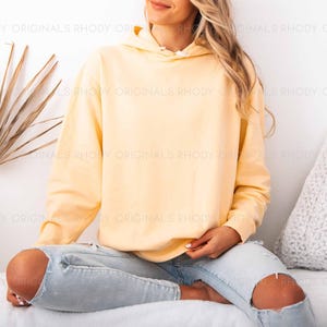 May include: A light yellow hoodie with a drawstring hood. The hoodie is worn with light blue ripped jeans. The background includes a white wall, a dried palm leaf, and a white pillow.