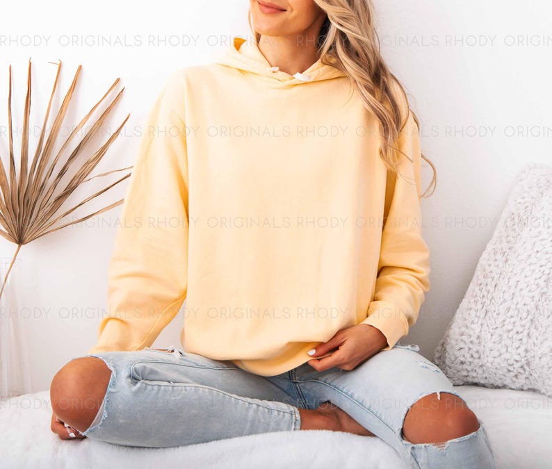 May include: A light yellow hoodie with a drawstring hood. The hoodie is worn with light blue ripped jeans. The background includes a white wall, a dried palm leaf, and a white pillow.