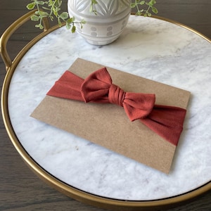 May include: A rust-colored baby headband with a bow, presented on a brown card. The headband is made of soft fabric and is displayed on a marble tray with a gold rim. A decorative vase with greenery is in the background.