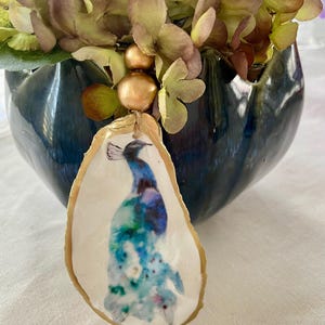 May include: An oyster shell ornament featuring a watercolor peacock design in shades of blue, green, and purple. The shell has a gold-painted edge and is suspended by a natural twine. The ornament is displayed in front of a dark blue vase with a floral arrangement.
