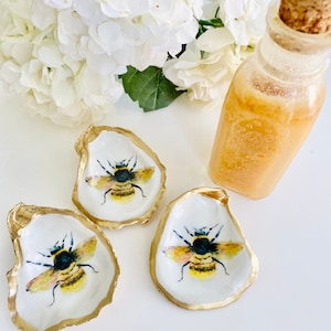 May include: Three small white oyster shell dishes with gold trim, each featuring a hand-painted bee with yellow and black stripes. The dishes are on a white surface next to a clear glass bottle with a cork stopper.
