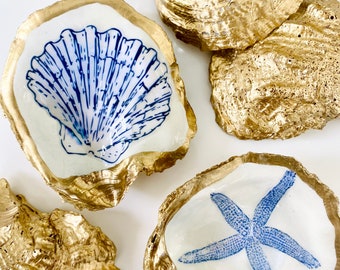 May include: Four gold oyster shells with white interiors. Each shell features a different blue and white nautical design: a seashell, a starfish, a seashell, and a starfish.