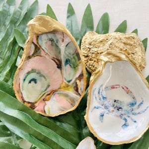 May include: Two gold-rimmed oyster shells, one with a pink and green marbled design and the other with a blue crab painted inside. The shells are on a bed of green leaves.