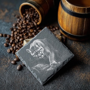 May include: A square, dark gray slate coaster featuring a detailed etched image of a cougar. The coaster is surrounded by coffee beans and wooden barrels, suggesting a coffee-themed product.