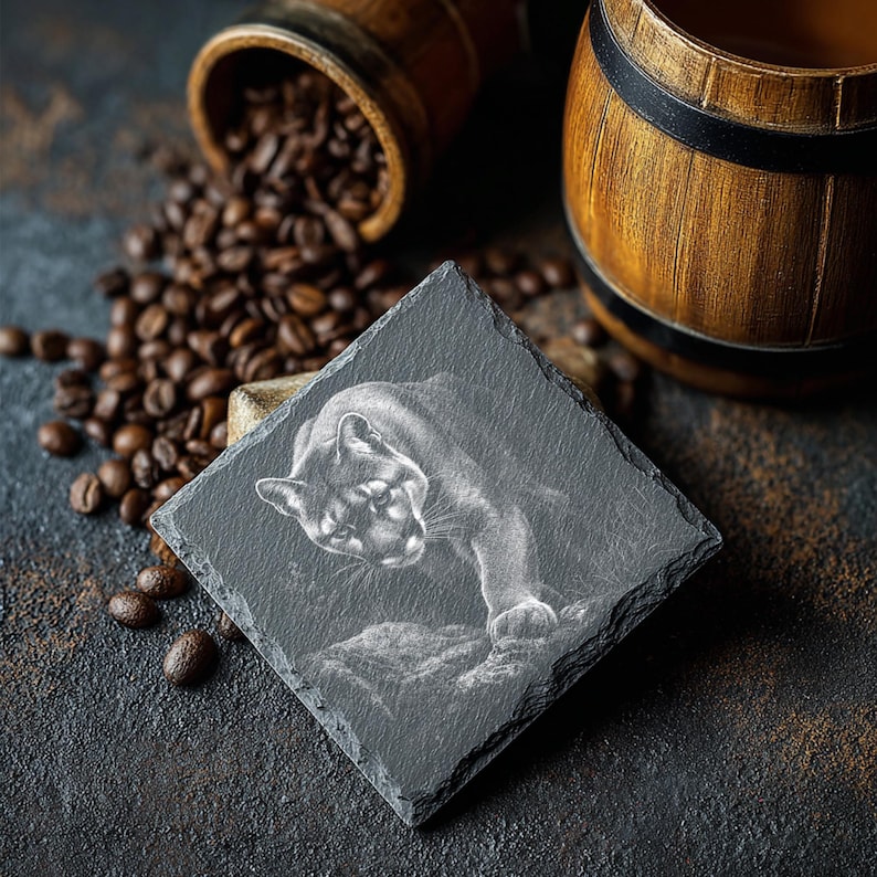 May include: A square, dark gray slate coaster featuring a detailed etched image of a cougar. The coaster is surrounded by coffee beans and wooden barrels, suggesting a coffee-themed product.