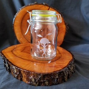 May include: A clear glass jar with a silver lid and a yellow rubber seal. The jar has an etched design of an alien with a UFO in the background.