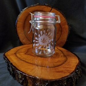 May include: A clear glass jar with a silver lid and a pink rubber seal. The jar has an etched design of a marijuana leaf inside a circle of water droplets. The jar is sitting on a wooden slab.