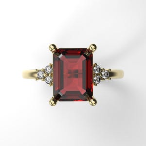 Peut inclure: Une bague en or ornée d'une grande pierre rouge foncé rectangulaire en son centre. De petits groupes de pierres blanches flanquent la pierre principale. La bague a un design simple et élégant, avec un anneau en or poli.
