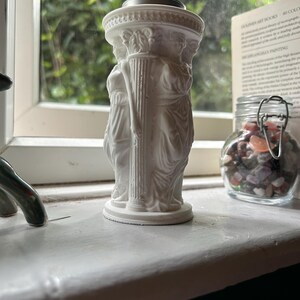 Könnte beinhalten: Ein weißer Kerzenhalter im klassischen Stil mit geformten Figuren und Säulen. Das detaillierte Design umfasst eine kreisförmige Ober- und Unterseite, wobei die Figuren den oberen Teil tragen. Der Kerzenhalter steht auf einer weißen Fensterbank.
