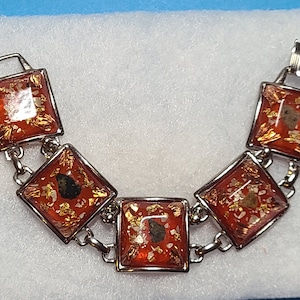 May include: A silver bracelet with five square-shaped red and gold resin charms. Each charm has a different pattern of gold flakes and black specks.