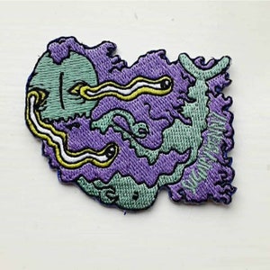 Op de afbeelding: Paarse en groene geborduurde patch met een cartoonoog met twee gele en witte wormen die eruit komen. De tekst "WORM" is geborduurd aan de rechterkant van de patch.