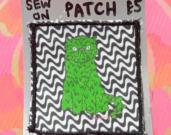 May include: A sew-on patch featuring a green, furry cat design with white eyes, set against a black and white wavy pattern. The patch is bordered with black stitching and includes the text "SEW ON PATCH ES".