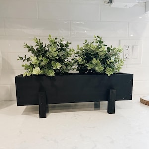 Könnte beinhalten: Eine schwarze, rechteckige Pflanzschale mit zwei Abschnitten mit grünen und hellgrünen künstlichen Eukalyptuspflanzen. Der Pflanzkübel wird von zwei schwarzen Stützen angehoben und steht auf einer weißen Arbeitsplatte.