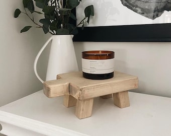 May include: A wooden stool with four legs shaped like a stylized animal. A brown glass candle with a white label is on top of the stool. The stool is on a white surface.