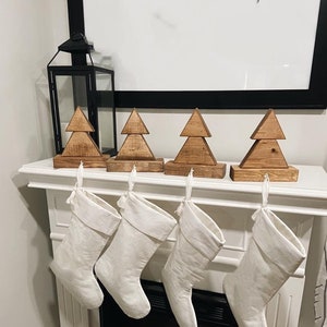 Peut inclure: Un manteau de cheminée blanc avec quatre bas de Noël blancs suspendus sous quatre décorations d'arbres de Noël en bois. Une lanterne noire est sur le côté gauche du manteau.