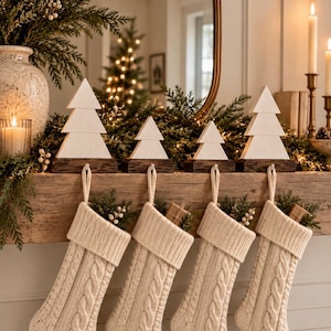 May include: Four cream-coloured cable knit stockings hang from a wooden mantel, each suspended by a white wooden Christmas tree stocking holder with a dark brown base. The stockings feature a textured knit pattern and are filled with greenery.