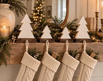 Puede incluir: Cuatro calcetines navideños de punto trenzado color crema cuelgan de una repisa de madera, cada uno sujeto por un soporte de árbol de Navidad blanco con base marrón oscuro. Los calcetines tienen un patrón de punto y contienen follaje.
