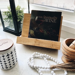 May include: A wooden book stand with a book titled "The Spirituality of Wine" by Tom Harpur. A white ceramic jar with a black and white geometric pattern, a white beaded garland with a brown tassel, and a wooden mortar and pestle are also in the image.